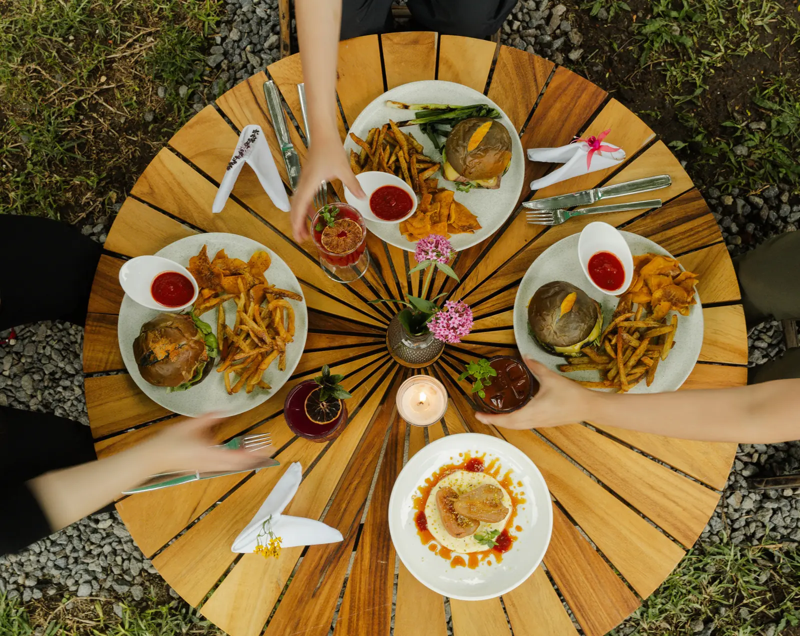 A shared table at Rosanta seen from above: burgers, fries, salsas, cocktails and flowers on a round wooden table